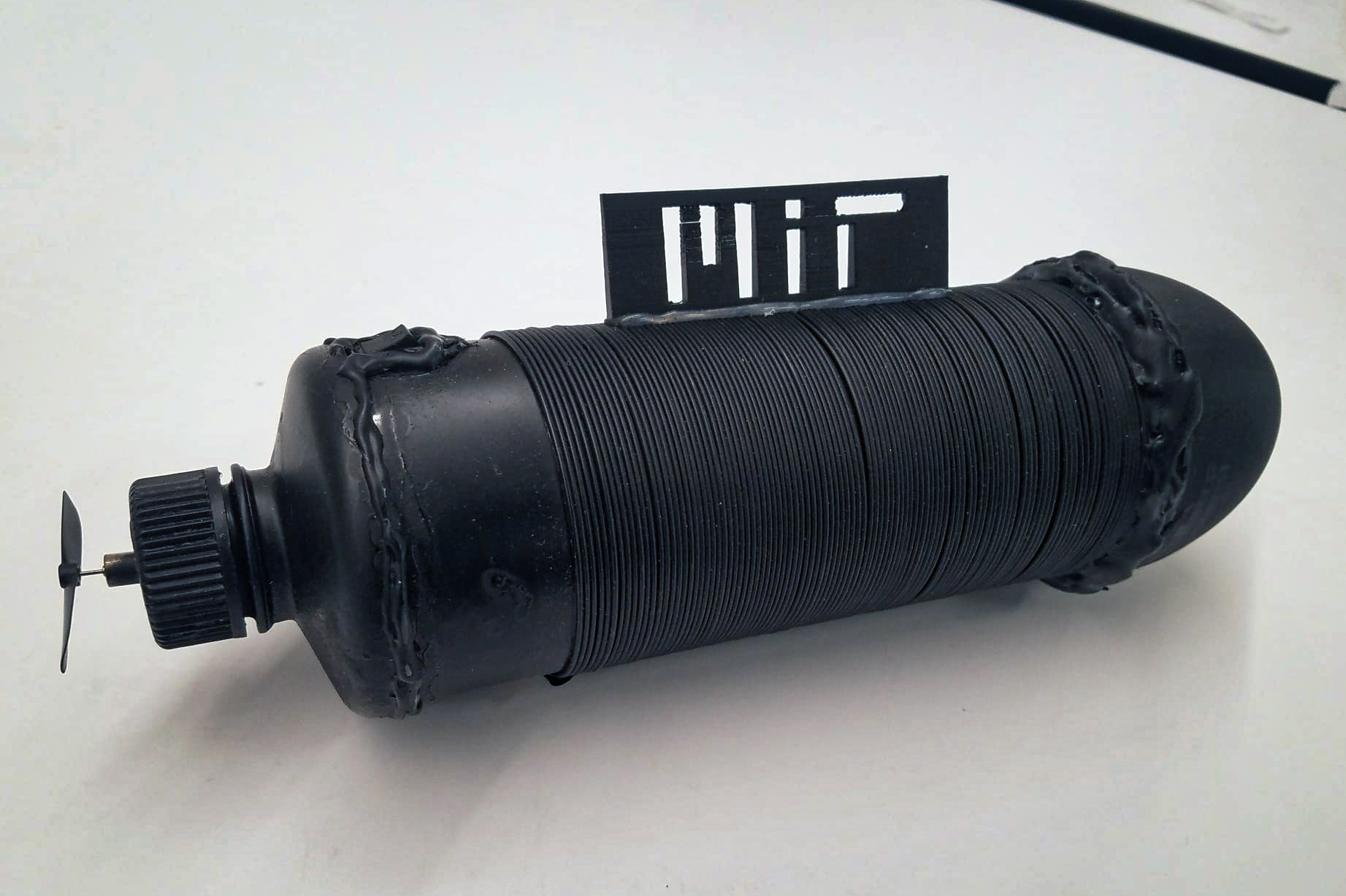 An oblong black tube, wrapped in a black wire, with an MIT logo on top  and a propeller at one end.