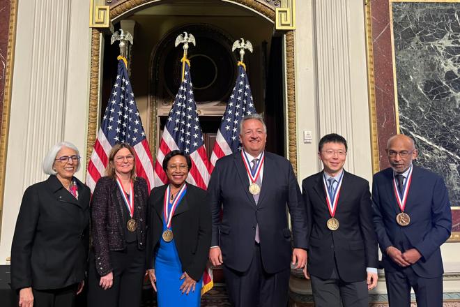 Six men and women stand in front of an array of American flags. Five of the six wear National Medals of Science or Technology and Innovation