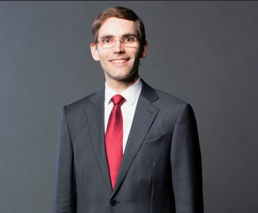 a man wearing a grey suit and red necktie stands in from of a grey backdrop