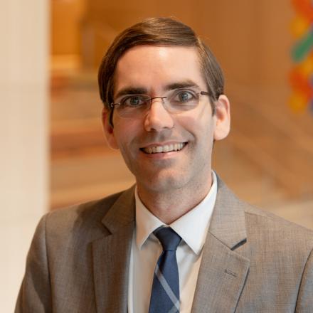 Portrait of MIT professor Tomás Palacios wearing glasses, a gray suit jacket, and a blue tie, smiling at the camera against a warm-toned background.