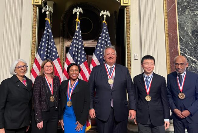 Six men and women stand in front of an array of American flags. Five of the six people are wearing National Medals of Science or National Medals of Technology and Innovation.