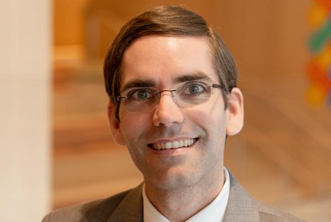 Professional headshot of Tomás Palacios wearing glasses, a light gray suit, white shirt, and blue striped tie, smiling directly at the camera against a softly blurred indoor background.