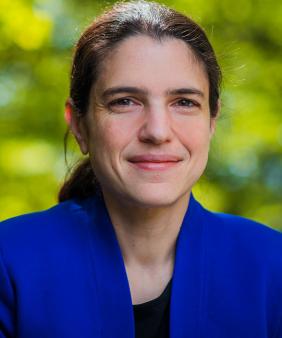 Prof. Heather J. Kulik, from the shoulders up, wearing a royal blue jacket over a black top, posed in front of a blurred, treed background