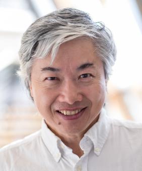 Dr. Shuguang Zhang from the shoulders up in front of a blurred background of pipes and beams